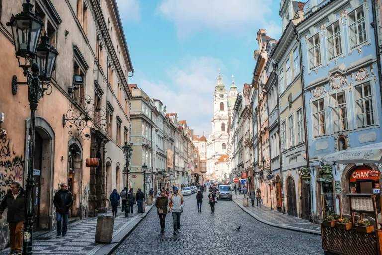A cobbled street in Prague's Malá Strana district, lined with historic townhouses and leading toward the baroque towers of St. Nicholas Church.